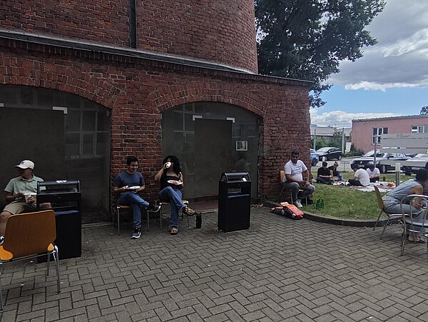 Mehrere Personen sitzen bei einem Sommerfest im Innenhof vor einem Backsteingebäude und essen gemeinsam.
