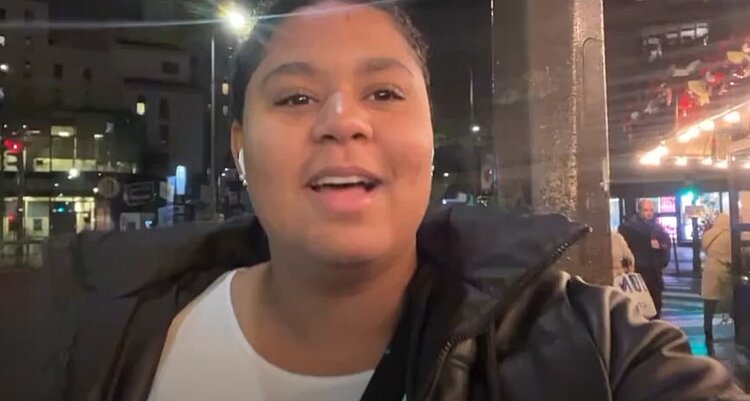 Eine junge Frau macht ein Video von sich. Sie hält dabei das Handy hoch und spricht in die Kamera. Sie hat weiße Airpods in den Ohren und trägt eine schwarze Winterjacke. Im Hintergrund sieht man einen Ausschnitt einer weihnachtlich beleuchteten Stadt.