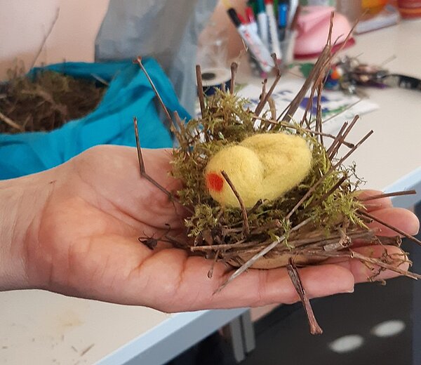 Einstimmung auf Ostern Auf der offenen Handfläche einer Auszubildenden liegt ein selbst gebasteltes Nest aus Moos und Stöckern, in dem ein selbst gestaltetes Küken aus Filz liegt.