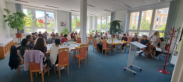 Speed-Dating für Pflegefachkräfte: Eine gelungene Vorbereitung für die Ausbildung Zu sehen ist ein großer Raum Raum mit einer weißen Säule in der Mitte. In dem Raum stehen überall in Vierecke zusammengestellte Tische mit Stühlen, auf denen jüngere mit mittelalte Personen sitzen.