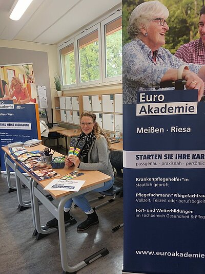 Eine Mitarbeitende der Euro Akademie mit lockigen Haaren und Brille sitzt an einem Tisch in einem Klassenraum. Sie trägt eine Strickjacke und hält mehrere bunte Fächer mit Informationsmaterial in der Hand. Auf dem Tisch liegen zahlreiche Broschüren und Flyer. Im Hintergrund stehen zwei große, blau-weiße Roll-ups mit dem Schriftzug 'Euro Akademie' und weiteren Informationen zu Aus- und Weiterbildungsmöglichkeiten im Pflegebereich. Durch die Fenster fällt Tageslicht, und an der Wand hängen Pinnwände mit Dokumenten.