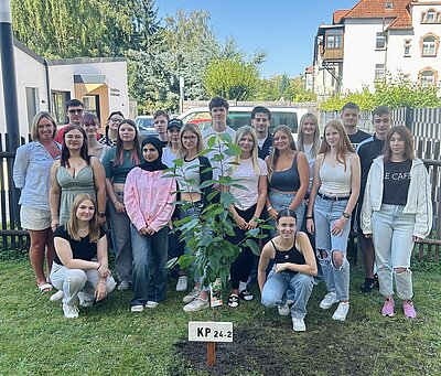 Eine Gruppe von Menschen steht im Freien um einen kleinen Baum. Sie posieren lächelnd für ein Gruppenfoto. Der Baum hat ein Schild mit der Aufschrift "KP 24-2". Die Gruppe besteht aus jungen Erwachsenen und sie stehen auf einem Rasen mit einem Gebäude im Hintergrund.