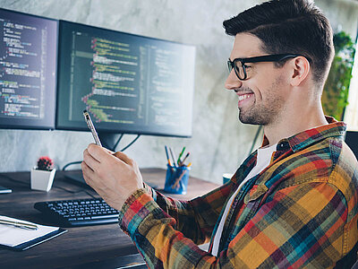 Ein junger Mann mit Brille sitzt lächelnd an einem Schreibtisch in einem modernen Büro. Er trägt ein kariertes Hemd in verschiedenen Farben und hält einen Stift in der Hand. Vor ihm stehen zwei große Bildschirme, auf denen Programmiercode zu sehen ist, was darauf hindeutet, dass er als Softwareentwickler oder Programmierer arbeitet. Auf dem Schreibtisch liegen außerdem eine Tastatur, ein Notizblock, ein Handy und ein paar Stifte. Daneben steht ein kleiner Topfpflanze. Der Mann wirkt zufrieden und konzentriert. Die Szene vermittelt eine produktive und kreative Arbeitsatmosphäre.