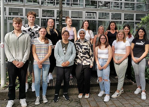 Herzlich willkommen Eine Gruppe von Jugendlichen posiert für ein Gruppenfoto. Die Mehrzahl sind Mädchen, es sind nur zwei Jungen auf dem Bild. Die Personen tragen sommerliche Kleidung und stehen in einem Hof vor einem Gebäude mit einer Glasfassade.