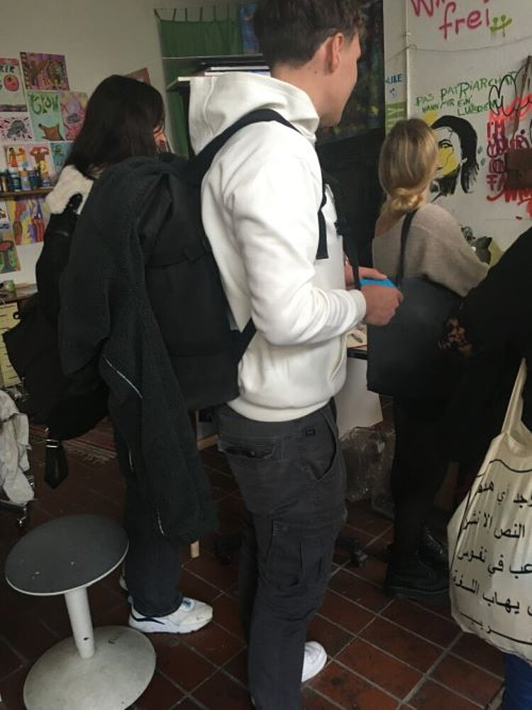 Mehrere Personen stehen in einem künstlerisch gestalteten Atelier und schauen sich bunte Graffitis und Zeichnungen an. Im Vordergrund steht ein Mann mit einem weißen Kapuzenpullover und einem schwarzen Rucksack.