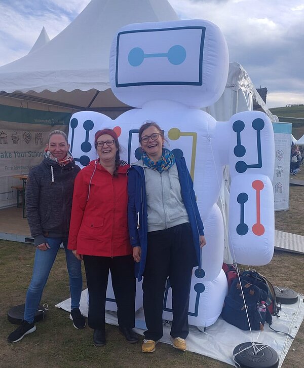 Drei lächelnde Personen stehen vor einem großen, aufblasbaren Roboter mit kindgerechtem Design auf einer Veranstaltung. Im Hintergrund ist ein Zelt mit einem Banner, auf dem „Make Your School“ und „Eure Ideenwerkstatt“ zu lesen ist.