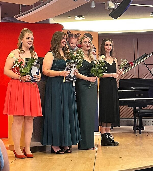 Feierlicher Abschied: Exmatrikulation der Absolvent*innen 2025 Eine Gruppe von Frauen steht auf einer Bühne und hält Blumen und Zertifikate. Sie sind elegant gekleidet und lächeln. Im Hintergrund ist ein rotes Banner und ein Klavier zu sehen.