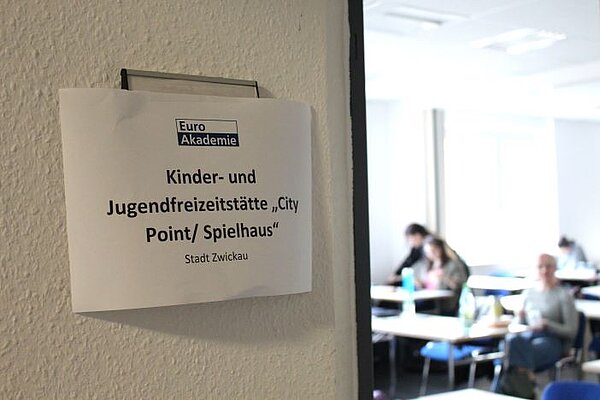 Theorie meets Praxis Vor einem Seminarraum ist ein Schild angebracht, auf dem das Logo der Euro Akademie sowie "Kinder- und Jugendfreizeitstätte City Point/Spielhaus" steht. Im Hintergrund ist unscharf der Raum mit Teilnehmenden, Tischen und Stühlen zu sehen.