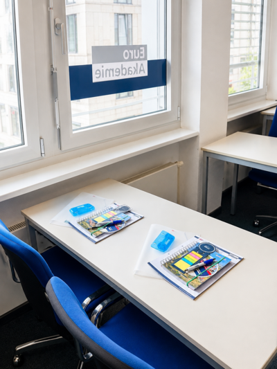 Ein Klassenzimmer mit weißen Tischen und blauen Stühlen. Auf den Tischen liegen Schreibutensilien, Notizblöcke und transparente Mappen. Durch das Fenster ist ein Schild mit der Aufschrift 'EURO Akademie' zu sehen. Die Fenster lassen Tageslicht herein und bieten einen Blick auf ein Gebäude im Hintergrund.