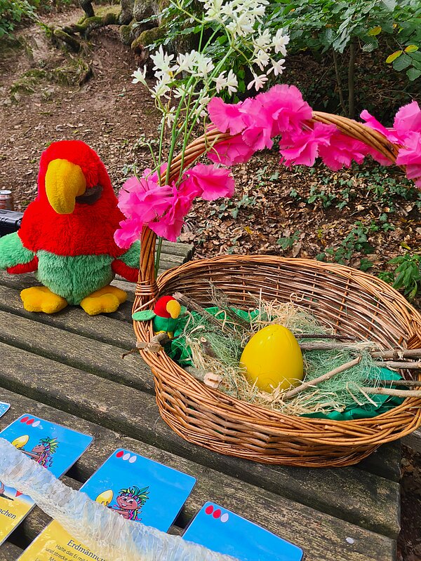 Kennenlerntag der Euro Akademie Rochlitz Ein Deko-Korb mit pinken Blumen und einem großen gelben Ei darin, daneben ein Plüschpapagei und Spielkarten auf einem Holztisch im Wald.