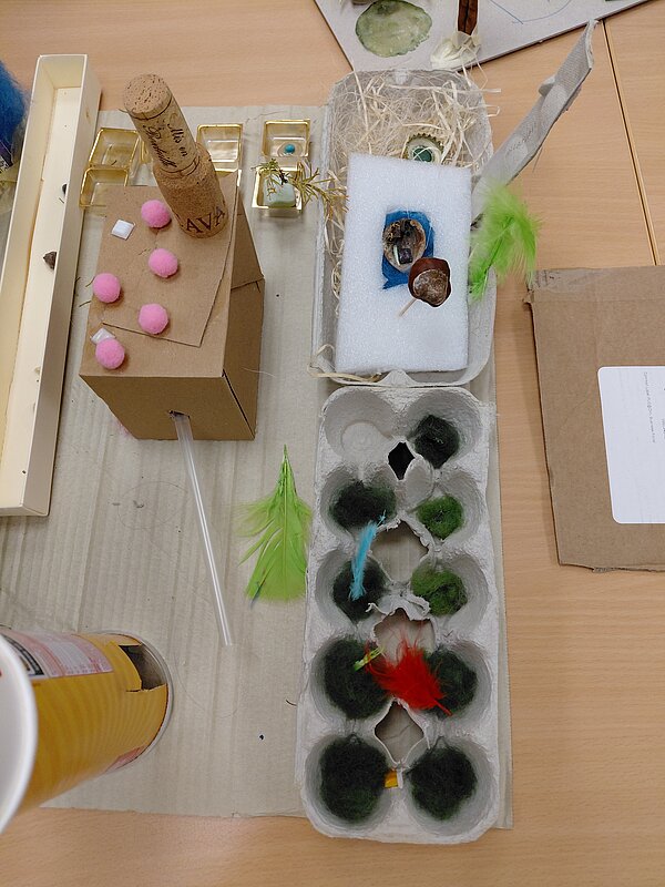 Auf einer großen Pappunterlage steht ein fantasievolles Kunstwerk aus Alltagsmaterialien: ein Eierkarton mit grüner Wolle und bunten Federn, daneben eine kleine Styroporplattform mit Kastanie und Muschel sowie ein aus Karton und Korken gebauter Turm mit rosa Pompons.