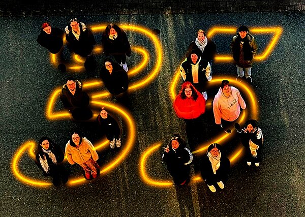 Mitarbeitende der Euro-Schulen und Euro Akademie Bitterfeld-Wolfen und Dessau stehen auf einer asphaltierten Fläche, auf der in leuchtendem Gelb die Zahl 35 groß aufgemalt ist, als Symbol zum 35-jährigen Jubiläum. Die Personen tragen überwiegend dunkle Kleidung, einige Jacken sind farbig. Die Szene ist aus der Vogelperspektive aufgenommen, sodass die Zahl und die Anordnung der Menschen klar erkennbar sind. Die Lichtreflexe der gelben Linien heben sich deutlich vom dunklen Untergrund ab.