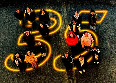 Mitarbeitende der Euro-Schulen und Euro Akademie Bitterfeld-Wolfen und Dessau stehen auf einer asphaltierten Fläche, auf der in leuchtendem Gelb die Zahl 35 groß aufgemalt ist, als Symbol zum 35-jährigen Jubiläum. Die Personen tragen überwiegend dunkle Kleidung, einige Jacken sind farbig. Die Szene ist aus der Vogelperspektive aufgenommen, sodass die Zahl und die Anordnung der Menschen klar erkennbar sind. Die Lichtreflexe der gelben Linien heben sich deutlich vom dunklen Untergrund ab.