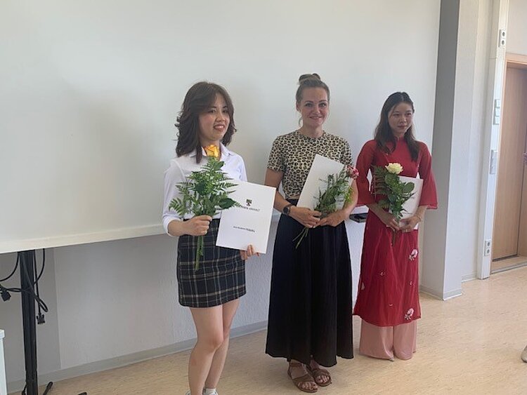 Herzlichen Glückwunsch! Drei junge Frauen stehen in einem hellen Raum vor einer weißen Projektionswand und halten ihre Abschlusszertifikate sowie Blumensträuße in den Händen. Sie lächeln und tragen verschiedene festliche Outfits – eine im weißen Hemd mit kariertem Rock, eine in einem gemusterten Oberteil mit schwarzem Rock und eine in einem roten Kleid mit Blumenmuster.