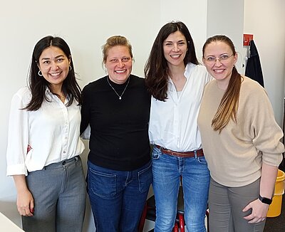 Vier erfolgreiche Absolventinnen einer Weiterbildung an der Euro Akademie Berlin stehen nebeneinander vor einer hellen Wand und posieren für ein Gruppenbild. Alle tragen langärmlige Oberteile in hellen oder neutralen Farben. Zwei Personen tragen weiße Hemden, eine trägt einen schwarzen Pullover, eine trägt ein beigefarbenes Oberteil. Zwei Personen tragen blaue Jeans, eine trägt eine graue Hose, eine trägt eine beigefarbene Hose. Im Hintergrund hängt ein dunkles Kleidungsstück an einem Haken. Die Gesichter sind unkenntlich gemacht.
