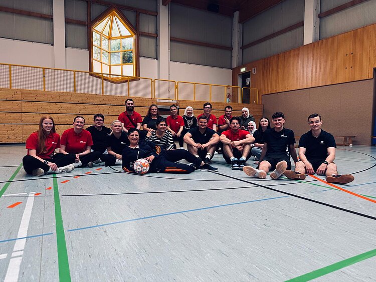 Gruppenfoto in einer Sporthalle: Mehrere Teilnehmende sitzen entspannt auf dem Hallenboden, einige tragen rote oder dunkle Shirts, im Vordergrund ist ein Fußball zu sehen.