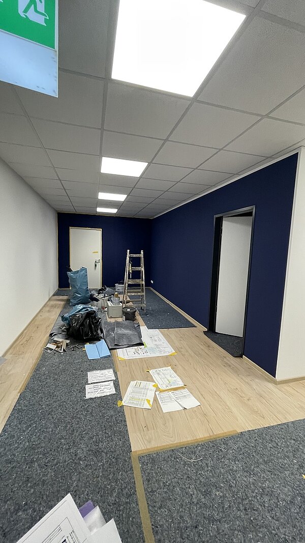 Blick in den Flur, in dem eine Wand dunkelblau gestrichen ist, Malerfließ ausliegt, eine Leiter aufgestellt ist und viele Papiere auf dem Boden liegen.
