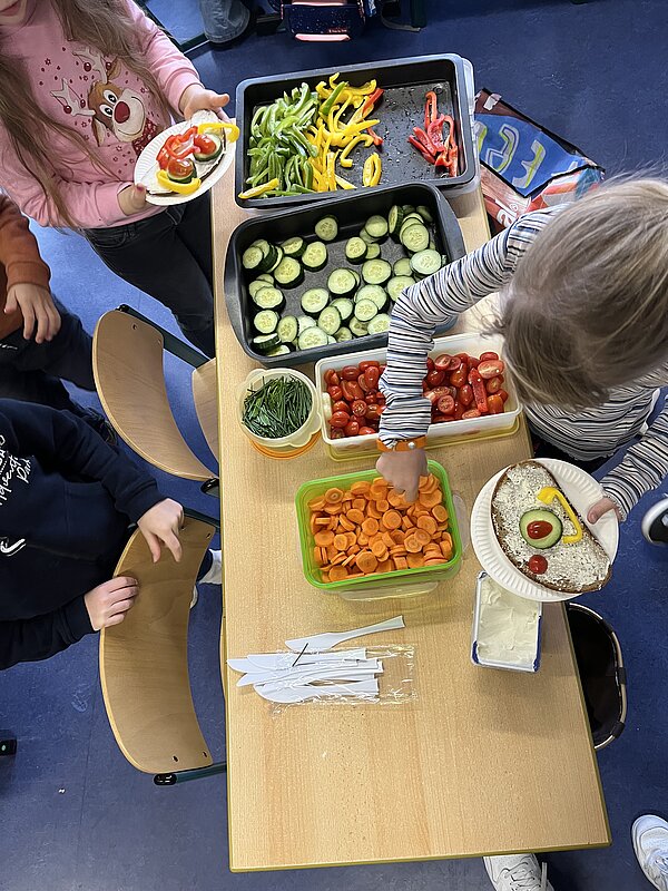 Aufsicht von oben auf einen Tisch mit geschnittenem Gemüse (Paprika, Gurken, Tomaten, Möhren) und Kräutern; Kinder belegen Brote auf Papptellern.