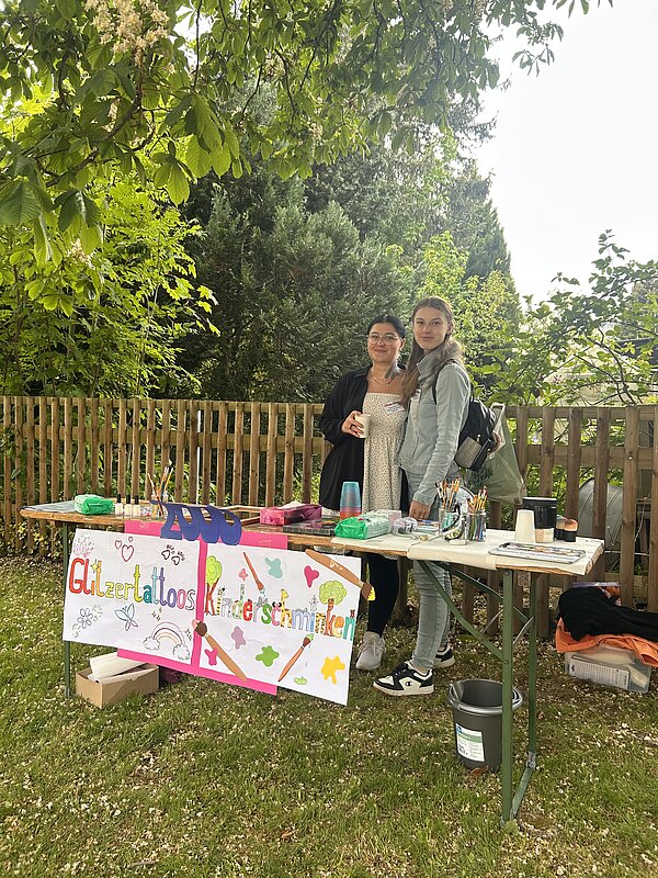 Zwei Personen stehen freundlich lächelnd hinter einem Tisch im Freien, auf dem ein bunt gestaltetes Schild steht, das auf Glitzertattoos und Kinderschminken hinweist. Der Tisch ist ausgestattet mit Farben, Pinseln und Zubehör für die Aktivitäten. Die Umgebung ist von viel Grün umgeben und wirkt freundlich und einladend.