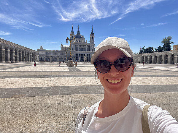 Erasmus-Bericht von Vanessa (Córdoba, 08/2024) Eine fröhliche Frau mit Sonnenbrille und Kappe lächelt in die Kamera, während sie auf dem weitläufigen Vorplatz des Königspalastes von Madrid steht. Im Hintergrund sind das prächtige Gebäude mit seinen Türmen sowie dekorative Laternen zu sehen. Der Platz ist mit hellem Stein gepflastert und einige Menschen spazieren darauf.