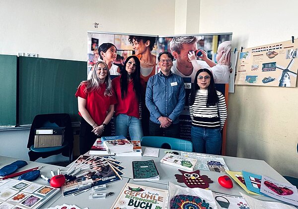 Vier lächelnde Personen stehen hinter einem Informationsstand in einem Klassenraum mit einer Tafel im Hintergrund. Drei von ihnen tragen rote oder gestreifte Oberteile, während eine Person ein blaues Hemd trägt. Auf dem Tisch vor ihnen liegen verschiedene Materialien wie Bücher, Broschüren, Stifte, Bastelutensilien und Informationsblätter. Im Hintergrund sind Poster mit Familienmotiven und pädagogischen Inhalten zu sehen.