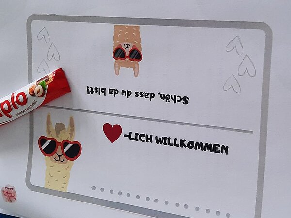 Ein Papierschild mit der Aufschrift 'Herzlich willkommen' und 'Schön, dass du da bist!' in verspielter Schrift. Daneben liegt ein Schokoladenriegel. Das Schild ist mit kleinen Illustrationen von Lama-Köpfen mit Sonnenbrillen verziert.