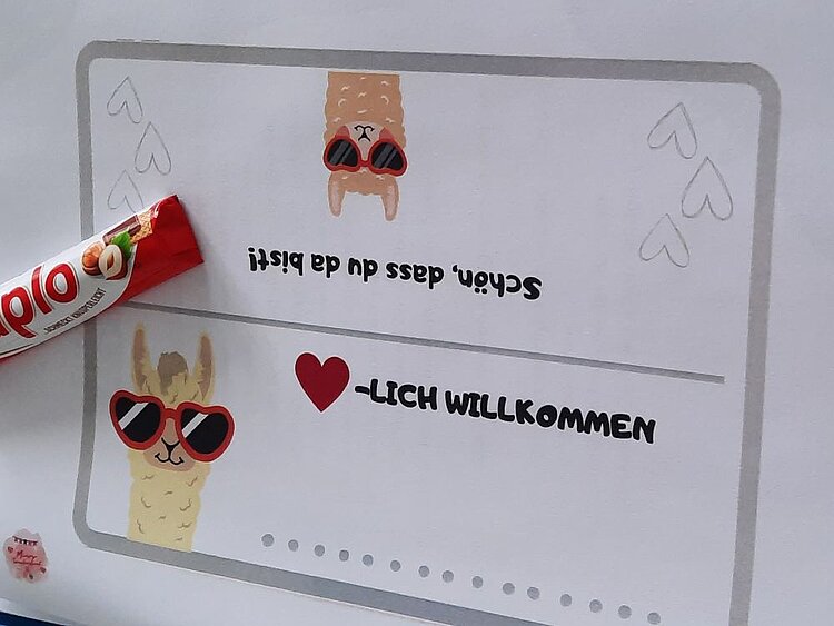 Ein Papierschild mit der Aufschrift 'Herzlich willkommen' und 'Schön, dass du da bist!' in verspielter Schrift. Daneben liegt ein Schokoladenriegel. Das Schild ist mit kleinen Illustrationen von Lama-Köpfen mit Sonnenbrillen verziert.