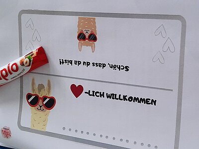 Ein Papierschild mit der Aufschrift 'Herzlich willkommen' und 'Schön, dass du da bist!' in verspielter Schrift. Daneben liegt ein Schokoladenriegel. Das Schild ist mit kleinen Illustrationen von Lama-Köpfen mit Sonnenbrillen verziert.