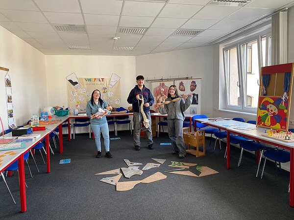 Drei Personen stehen in einem Klassenzimmer, das mit verschiedenen Lehrmaterialien ausgestattet ist. Eine Person hält ein Baby-Puppenmodell, eine andere hält ein Modell der Wirbelsäule, und die dritte Person hält ein Herzmodell. Im Hintergrund sind Tische, Stühle und ein Puppentheater zu sehen.