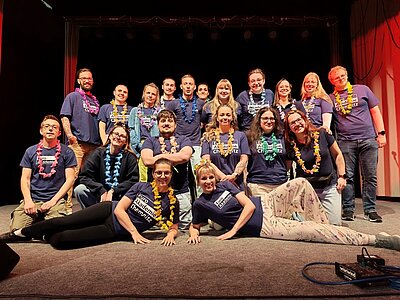 Gruppenbild der Auszubildenden auf der Bühne, alle tragen bunte Blumenketten um den Hals und überwiegend dunkelblaue T-Shirts. Einige Personen stehen, andere sitzen oder liegen auf dem Boden. Im Hintergrund sind Scheinwerfer und ein roter Vorhang zu sehen. Die Gesichter sind unkenntlich gemacht.