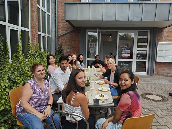 Eine Gruppe sitzt an zusammengestellten Tischen im Innenhof vor dem Eingang der Euro Akademie und Euro-Schulen.