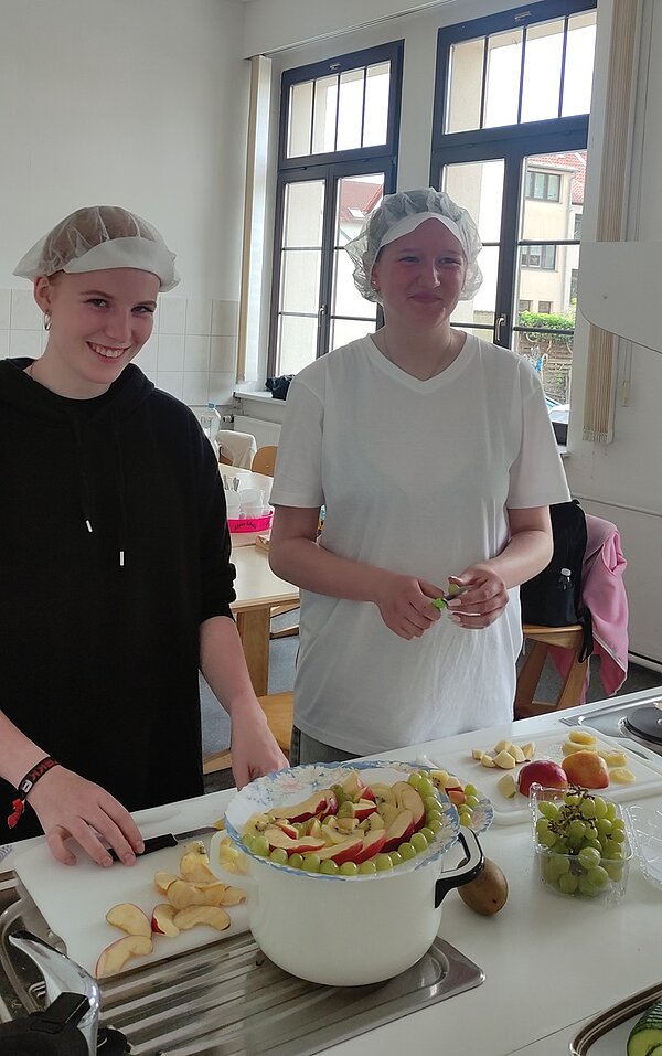 Gesund und kreativ Zwei angehende Kinderpflegerinnen stehen in der Küche und bereiten gesunde und bunte Snacks aus Obst und Gemüse vor. Sie tragen Hygienehauben auf dem Kopf und posieren lächelnd für ein Foto.