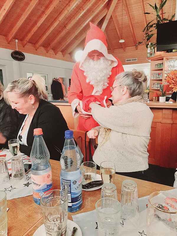 Nahaufnahme: Nikolaus im roten Kostüm überreicht etwas / spricht mit einer Frau am Tisch; viele Gläser und Wasserflaschen im Vordergrund.