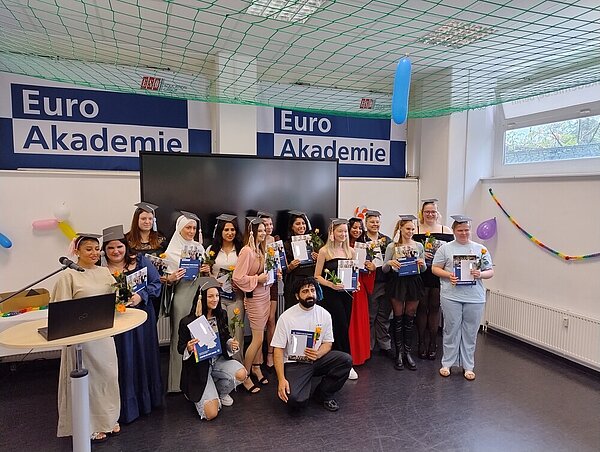 Eine Schülergruppe in festlicher Kleidung mit Absolventenhüten und gelben Rosen in der Hand hat sich für ein Foto positioniert. An der Wand hängen zwei große Logos der Euro Akademie, die Wände sind mit Luftballons und Girlanden feierlich geschmückt. Von der Decke hängt ein grünes Netz.
