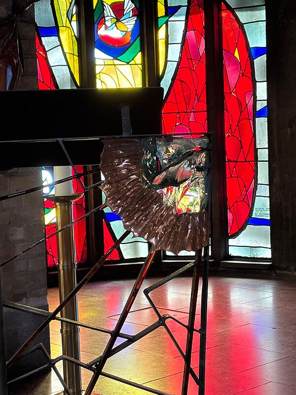 Detailaufnahme eines Metallgeländers mit kunstvollem Muster vor buntem Kirchenfenster mit abstrakten roten, gelben und blauen Glasmalereien.