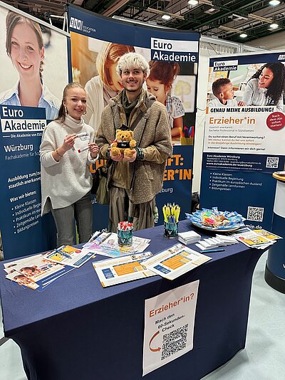 Zwei Personen stehen an einem Messestand der Euro Akademie vor großen blauen und weißen Infowänden, halten einen kleinen Teddybären mit Doktorhut und einen Giveaway-Stift in der Hand.