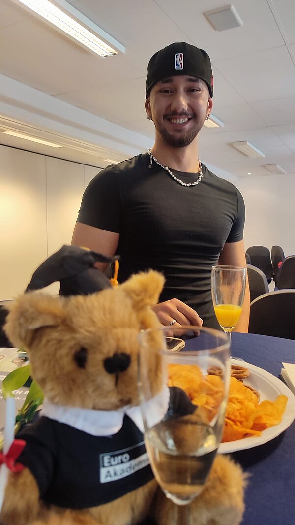 Ein mit einem Euro Akademie bekleideter Teddybär sitzt auf einem Tisch, dahinter steht ein junger man mit schwarzem Shirt und schwarzer Kappe. 