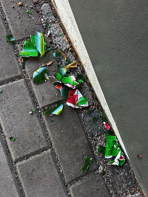 Eine zerbrochene grüne Glasflasche liegt am Bordstein auf der Straße.