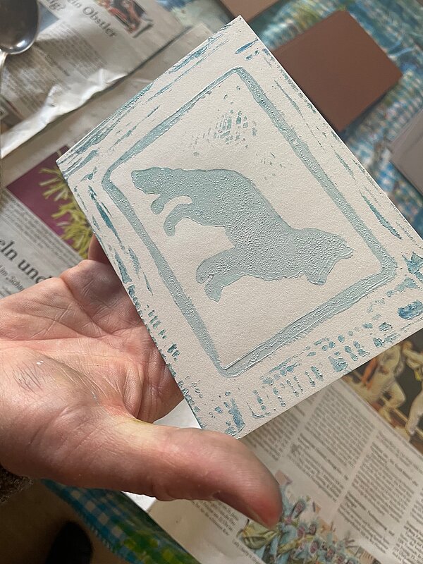 Eine Hand hält eine kleine Druckplatte mit dem Motiv eines springenden Tieres in hellblauer Farbe vor einem mit Zeitungen bedeckten Tisch.