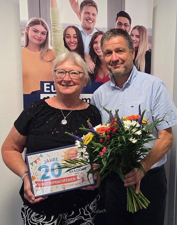 Sprachferien der Euro Akademie Jena und Erfurt: eine 20-jährige Erfolgsgeschichte Eine Frau und ein Mann stehen lächelnd vor einem Roll-up und blicken in die Kamera. In den Händen halten sie einen Bilderrahmen und einen Blumenstrauß.
