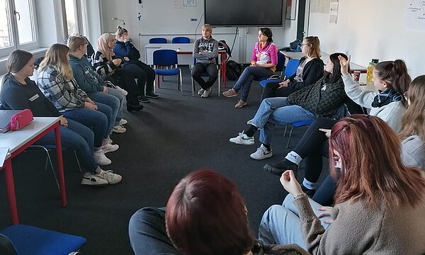 Sozialassistent*innen vertiefen Wissen über Cybermobbing Eine Gruppe von Schülern und Schülerinnen sitzt in einem Stuhlkreis in einem Klassenzimmer. Eine Schülerin hebt die Hand, um zu signalisieren, dass sie sprechen möchte. Es scheint eine lebhafte Diskussion zum Thema (Cyber)Mobbing zu laufen.