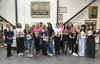 Gruppenfoto von jungen Erwachsenen und einer Lehrkraft, die lächelnd vor einer Wand stehen, an der ein Gemälde und historische Fotos hängen. Die Personen halten Zertifikate und kleine Geschenke in den Händen. 