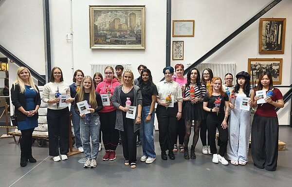 Herzlicher Abschied, freudiger Anfang: Pflege-Absolvent*innen geehrt und neue Auszubildende willkommen geheißen Gruppenfoto von jungen Erwachsenen und einer Lehrkraft, die lächelnd vor einer Wand stehen, an der ein Gemälde und historische Fotos hängen. Die Personen halten Zertifikate und kleine Geschenke in den Händen.