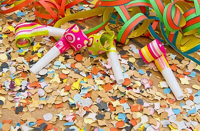 Drei bunte Party-Tröten in Pink, Grün und Gelb mit Streifen und Mustern liegen auf einem Tisch zwischen Konfetti und bunten Luftschlangen in verschiedenen Farben.