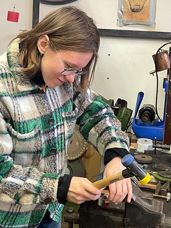 Eine Person mit Brille und kariertem Oberteil bearbeitet mit einem Hammer ein kleines Metallteil in einer Halterung. Im Hintergrund Werkstattgeräte und eine Maschine.