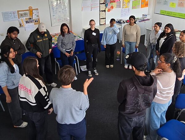 Teilnehmende stehen in einem Klassenraum im Kreis und diskutieren gemeinsam während eines Workshops.