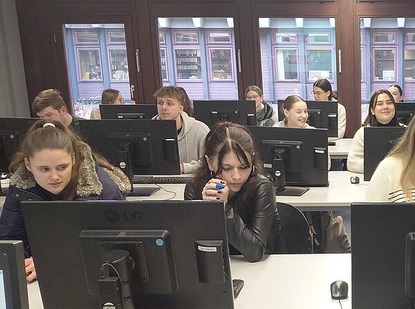 In einem hellen Computerraum der Universitätsbibliothek Würzburg sitzen mehrere angehende Erzieher der Euro Akademie Tauberbischofsheim an langen weißen Tischen vor schwarzen LG-Computermonitoren. Die Personen sind in lockerer Kleidung, einige tragen Pullover oder Jacken. Im Hintergrund sind große Fenster mit Blick auf eine Fassade mit mehreren Fenstern zu sehen, die den Raum mit Tageslicht erhellen. Die Anordnung der Tische und Monitore ist in Reihen, die Personen konzentrieren sich auf die Bildschirme oder blicken nach vorne. Die Atmosphäre wirkt ruhig und lernorientiert.