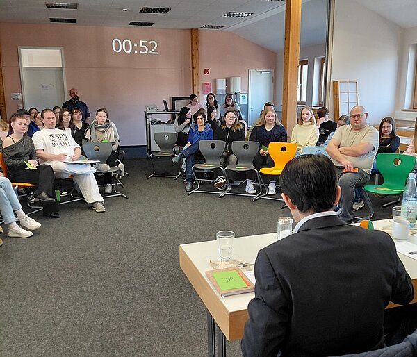 Großer Seminarraum der Euro Akademie Tauberbischofsheim mit zahlreichen sitzenden Studierenden und Mitarbeitenden, die aufmerksam einer Diskussionsrunde folgen. Im Vordergrund ist der Rücken eines sitzenden Politikers zu sehen, der an einem Tisch mit Wasserflaschen, Gläsern und Unterlagen Platz genommen hat. Die Teilnehmenden sitzen auf unterschiedlich farbigen Stühlen in mehreren Reihen und blicken konzentriert nach vorne. An der Wand ist eine digitale Zeitanzeige zu erkennen. Das Foto entstand während eines Besuchs zweier Politiker an der Euro Akademie Tauberbischofsheim und zeigt einen offenen Austausch zu bildungspolitischen und gesellschaftlichen Themen in akademischer Atmosphäre.