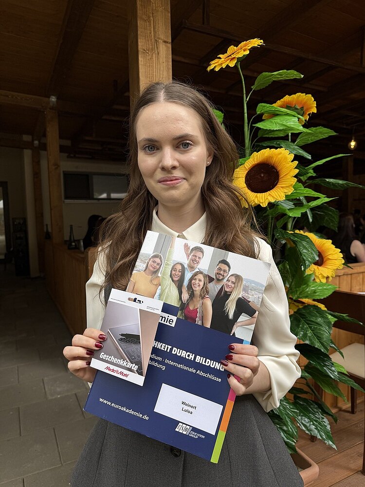 Erfolgreiche Graduation-Feier der Euro Akademie Bamberg/Nürnberg 2025 Luisa Weinert wurde als beste Absolventin geehrt und hält auf dem Foto Ihr Abschlusszeugnis sowie die Geschenkkarte von Media Markt in die Kamera. Sie trägt eine beigefarbene Bluse und einen grauen Rock, ihre Fingernägel sind dunkelrot lackiert.