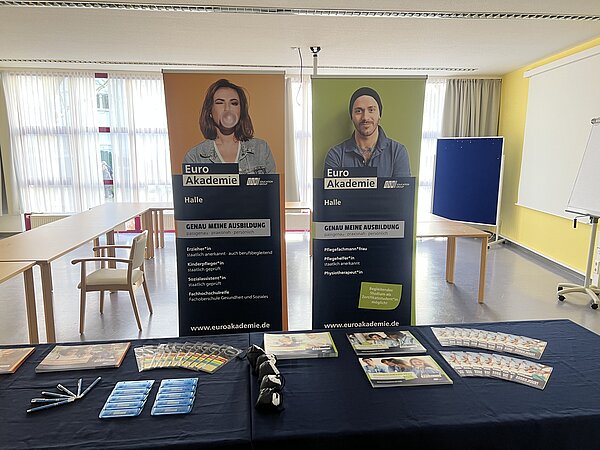 Informationsstand der Euro Akademie in einem hellen Raum: Auf einem dunkelblauen Tisch liegen Broschüren, Stifte und Give-aways vor zwei großen Roll-up-Bannern mit Ausbildungsangeboten.